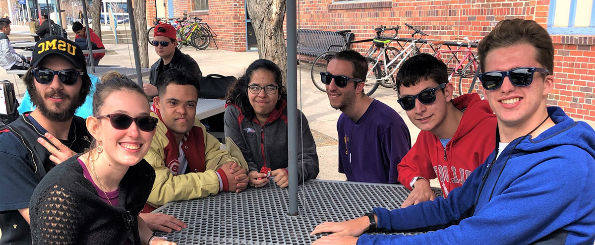 IHES students seated around an outdoor table, most wearing sunglasses and smiling for the camera.